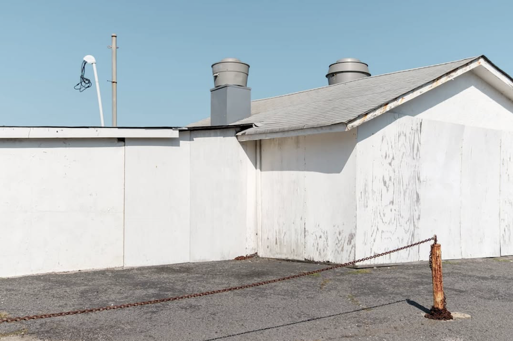A stark white industrial building with rooftop vents stands behind a rusted post and sagging chain under a clear blue sky. ©CynthiaGladis