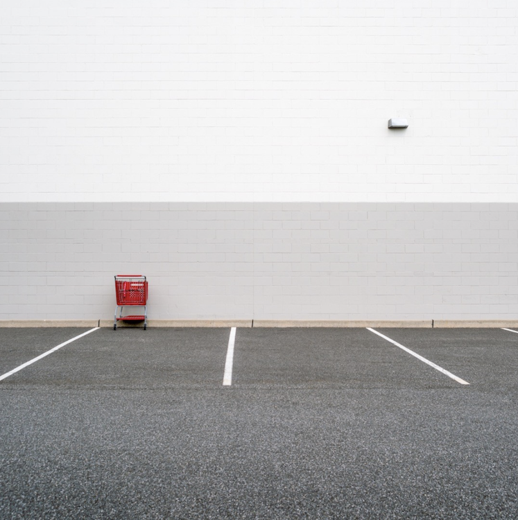 A lone red shopping cart sits in an empty marked parking space before an expansive white wall. ©CynthiaGladis
