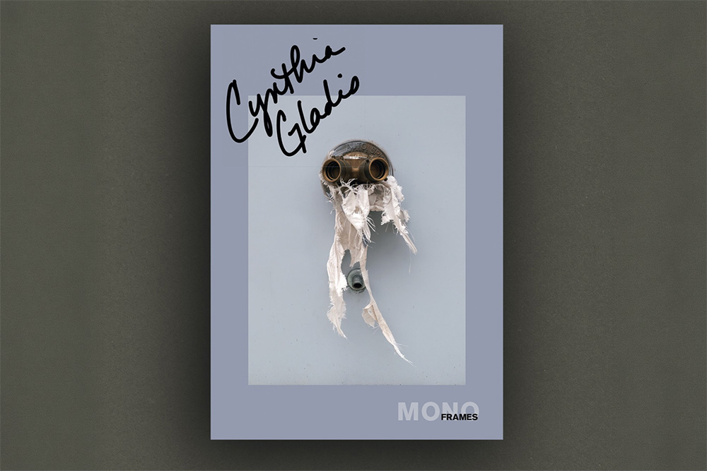 A gray MONO FRAMES publication cover bearing the handwritten name “Cynthia Gladis” features a surreal, mask-like assemblage of metal and torn white fabric. ©FramesMagazine for photo of book; ©CynthiaGladis for cover image