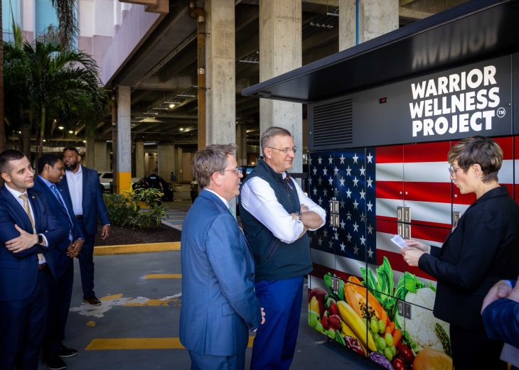 Several people stand in an outdoor area near a structure labeled “Warrior Wellness Project™,” which features an American flag design and images of food on its lockers.