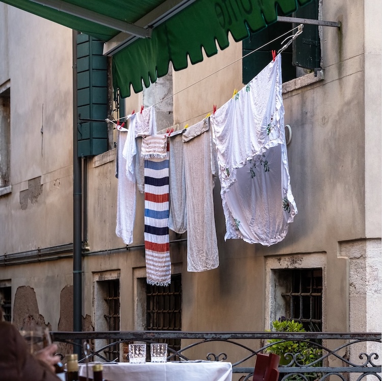 White garments and a striped towel hang above café tables outside a weathered building with dark green shutters and awning. ©CynthiaGladis