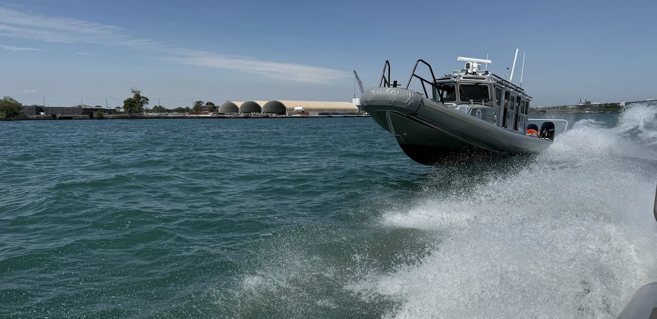 A grey roofed boat is shown bumping over waves in a river, with sea spray coming off another passing boat.