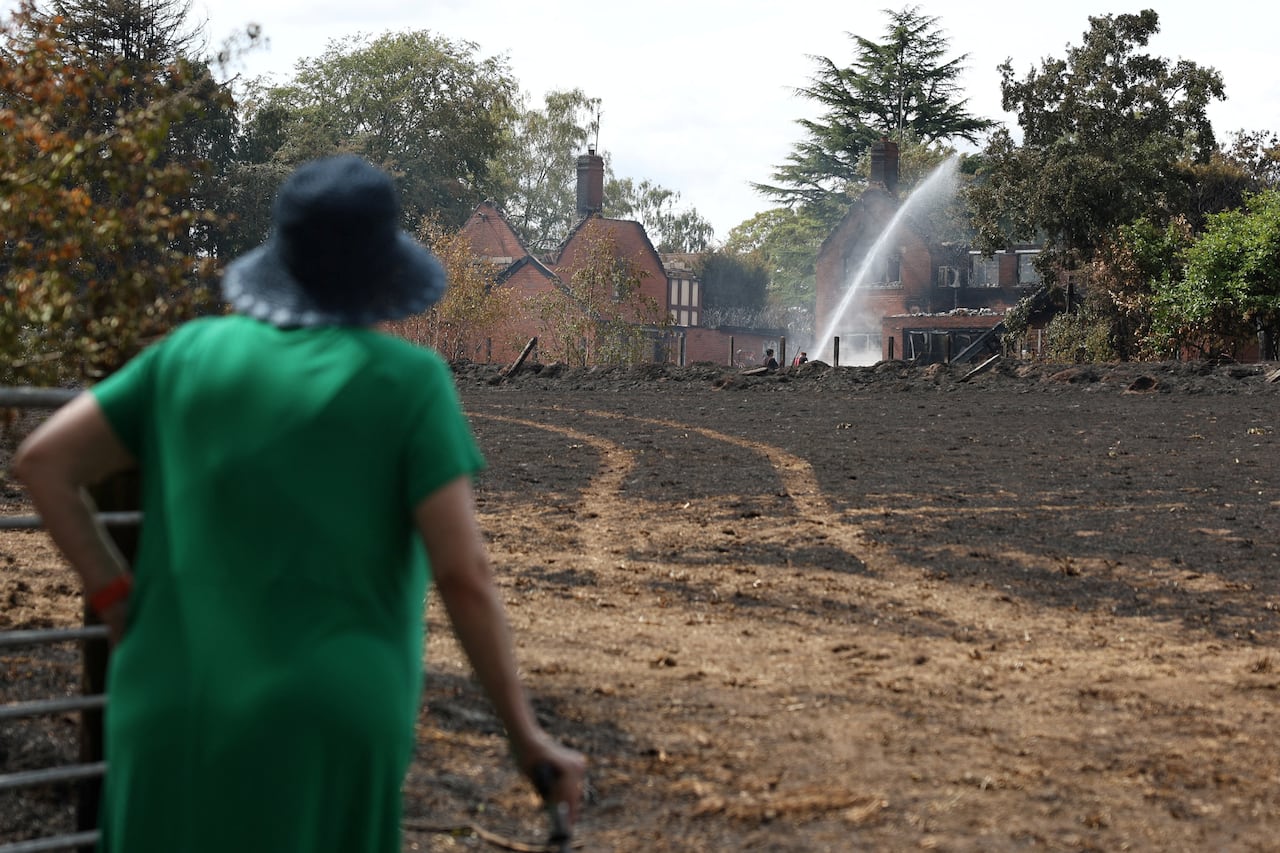 A woman in a green dress and blue hat watches a burnt house get sprayed with water.