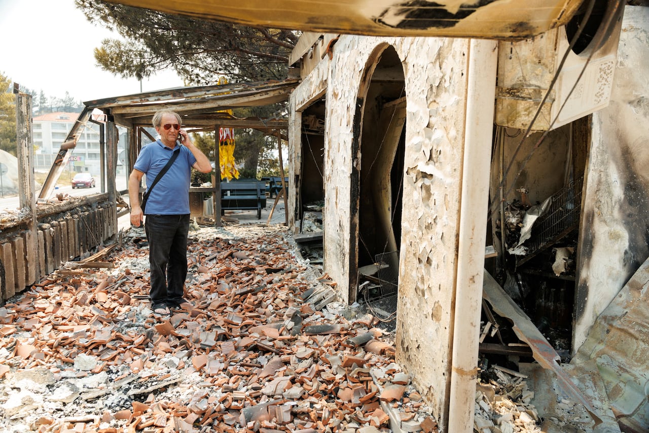 A man in a blue polo and black pants talks on the phone as he observes a badly burnt and damaged building.