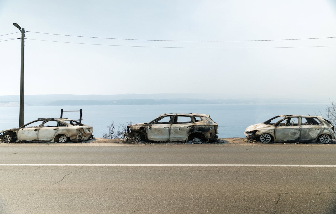 Three burnt cars are seen parked on a road in front of the sea.