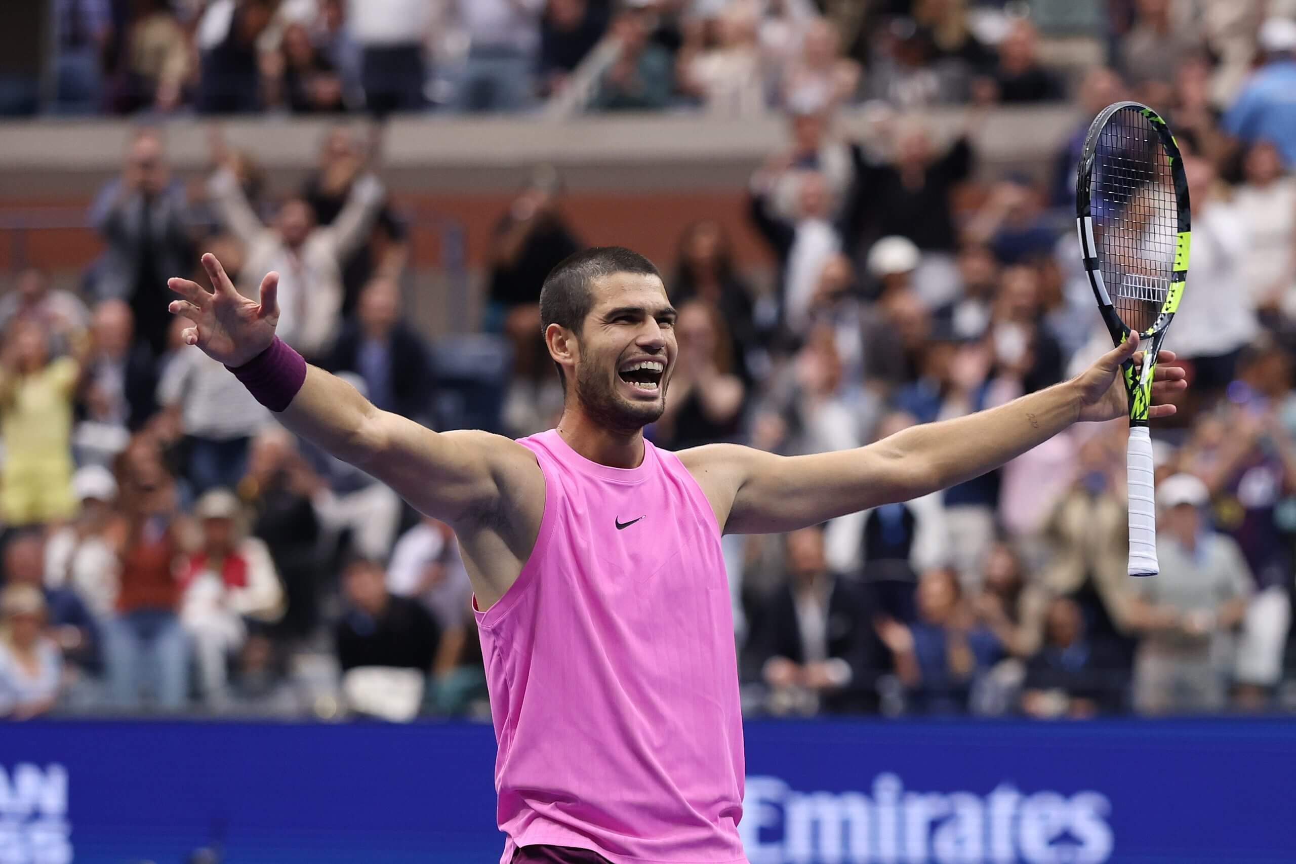 Carlos Alcaraz raises his racket with a wide grin wearing a pink vest.
