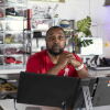 Richard Brown runs Proof Culture, a sneaker accessory company, out of his Ohio home. He is Black and has a beard. He is seated behind a computer monitor and there are sneakers in plastic display cases behind him.