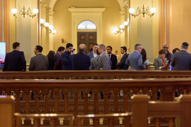 People wearing suits and nice clothing stand and talk amongst each other in a room with a hallway that has tall ceilings. A few wooden bars separate the people from the foreground.