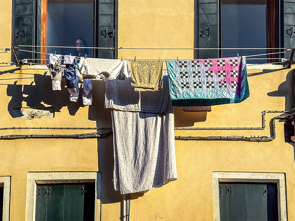 Colorful laundry hangs across the upper windows of a sunlit yellow Italian façade, casting crisp shadows on the wall. ©CynthiaGladis