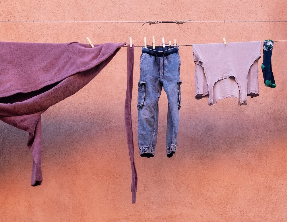 Purple fabric, jeans, a pale shirt, and a single sock hang from a clothesline against a salmon-pink stucco wall. ©CynthiaGladis