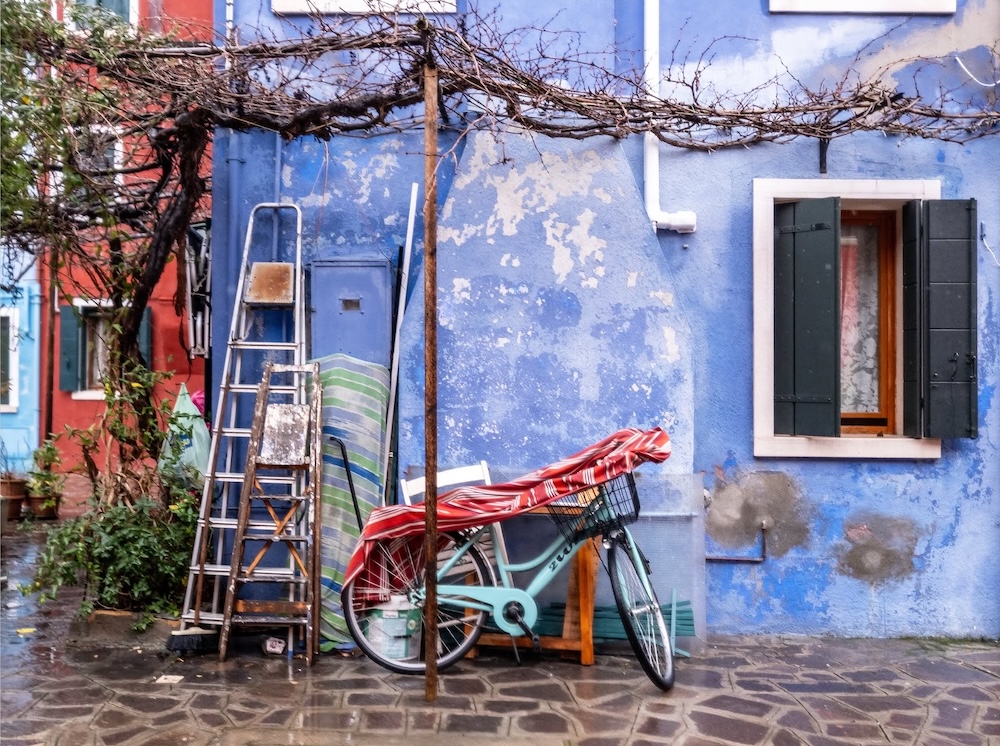 A bicycle, ladder, folded chairs, and bare grapevine cluster against a weathered blue house with dark shutters. ©CynthiaGladis