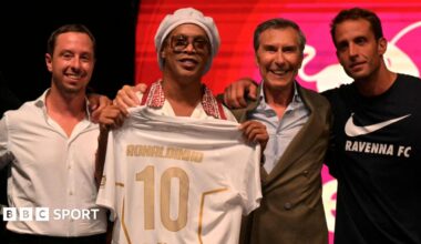 Ronaldinho holds a shirt bearing his name at a presentation event, he has one man to his right and two to his left