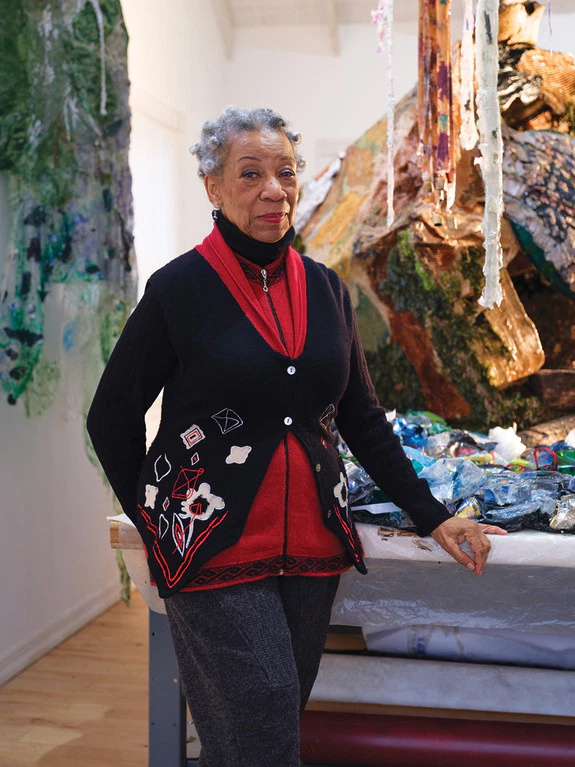 Smiling woman in a cozy sweater stands in an art studio, surrounded by colorful materials and abstract artwork.