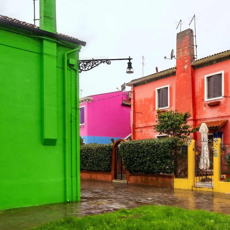 Vivid green, pink, orange, and yellow houses border a rain-darkened walkway and bright strip of grass beneath a pale sky. ©CynthiaGladis