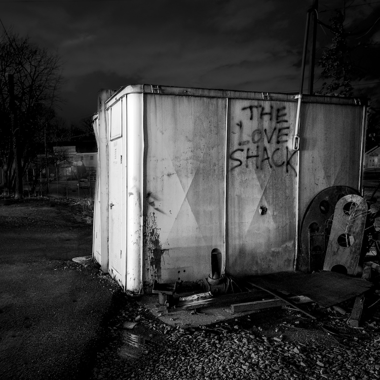 In a black-and-white night scene, a battered shed marked “THE LOVE SHACK” stands beside railroad tracks beneath a brooding sky. ©CynthiaGladis
