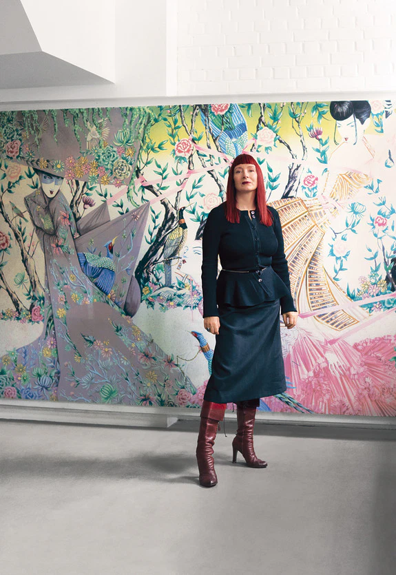 Person with red hair stands confidently in front of a colorful floral mural, wearing a dark outfit and red boots.