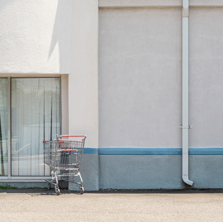 A shopping cart rests beside the glass door and drainpipe of a pale building with a blue-gray base. ©CynthiaGladis