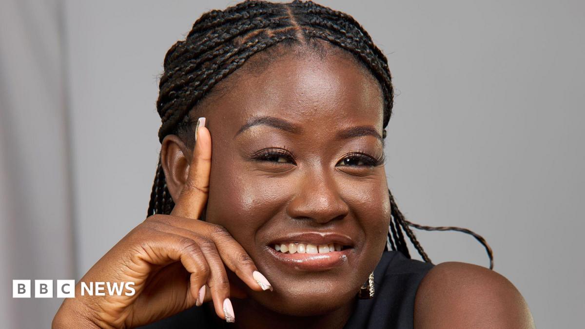 A woman with braids is smiling and leaning her face on her hand. She is wearing a black top and is photographed in front of a grey background.