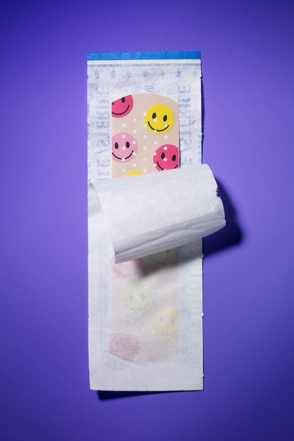 Colorful band-aid featuring smiley face designs on a purple background, partially unwrapped from its packaging.