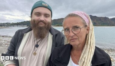 Ryan Shields and his mother Aly Shields standing together looking towards the camera. His mother is wearing a pink bandana on her blonde hair and has brownish glasses. Ryan is wearing greyish jacket and a blue beanie. He also has a long, light brown beard.