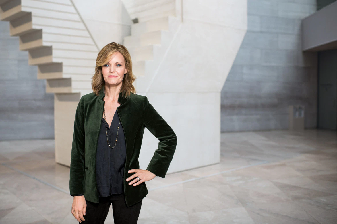 Person standing confidently in a modern building with gray walls and staircase in the background.