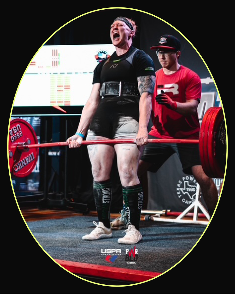 Athlete performing a deadlift with visible effort during a powerlifting competition, assisted by a teammate.