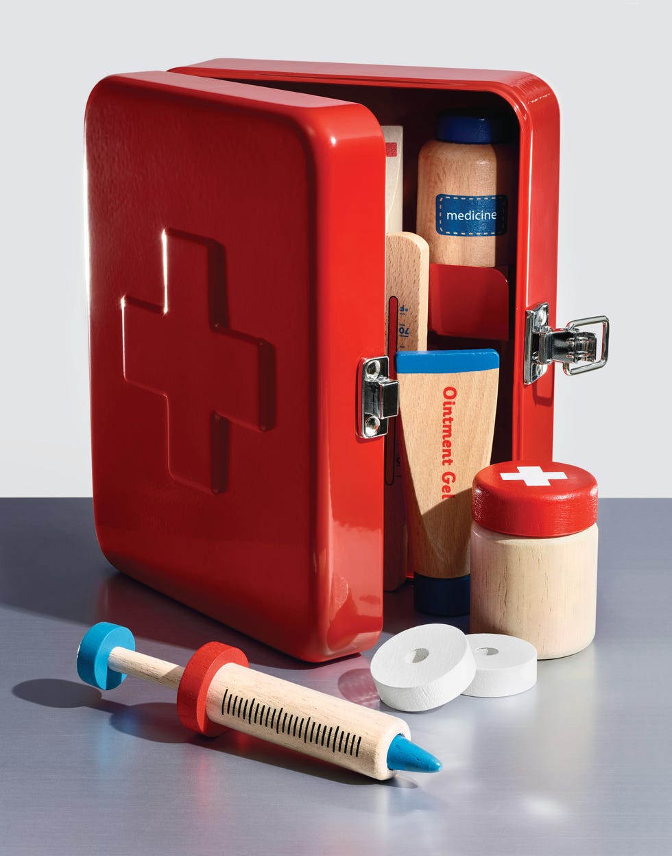 Toy red first aid kit with wooden medical supplies including tubes, a bottle, and a syringe on a silver surface.