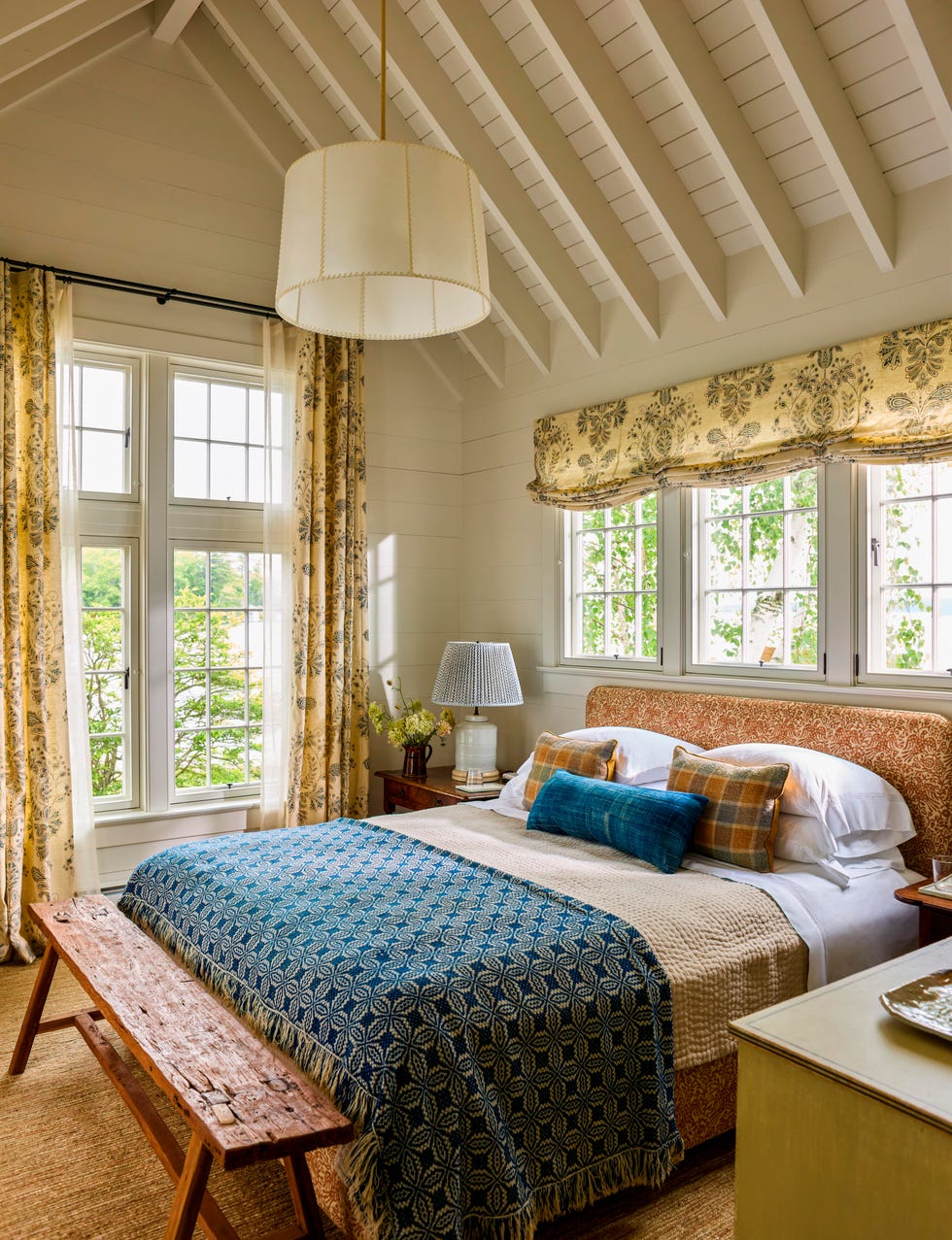 A bright, cozy bedroom with a patterned blanket, soft pillows, and large windows featuring floral curtains. A bright, cozy bedroom with a patterned blanket, soft pillows, and large windows featuring floral curtains.