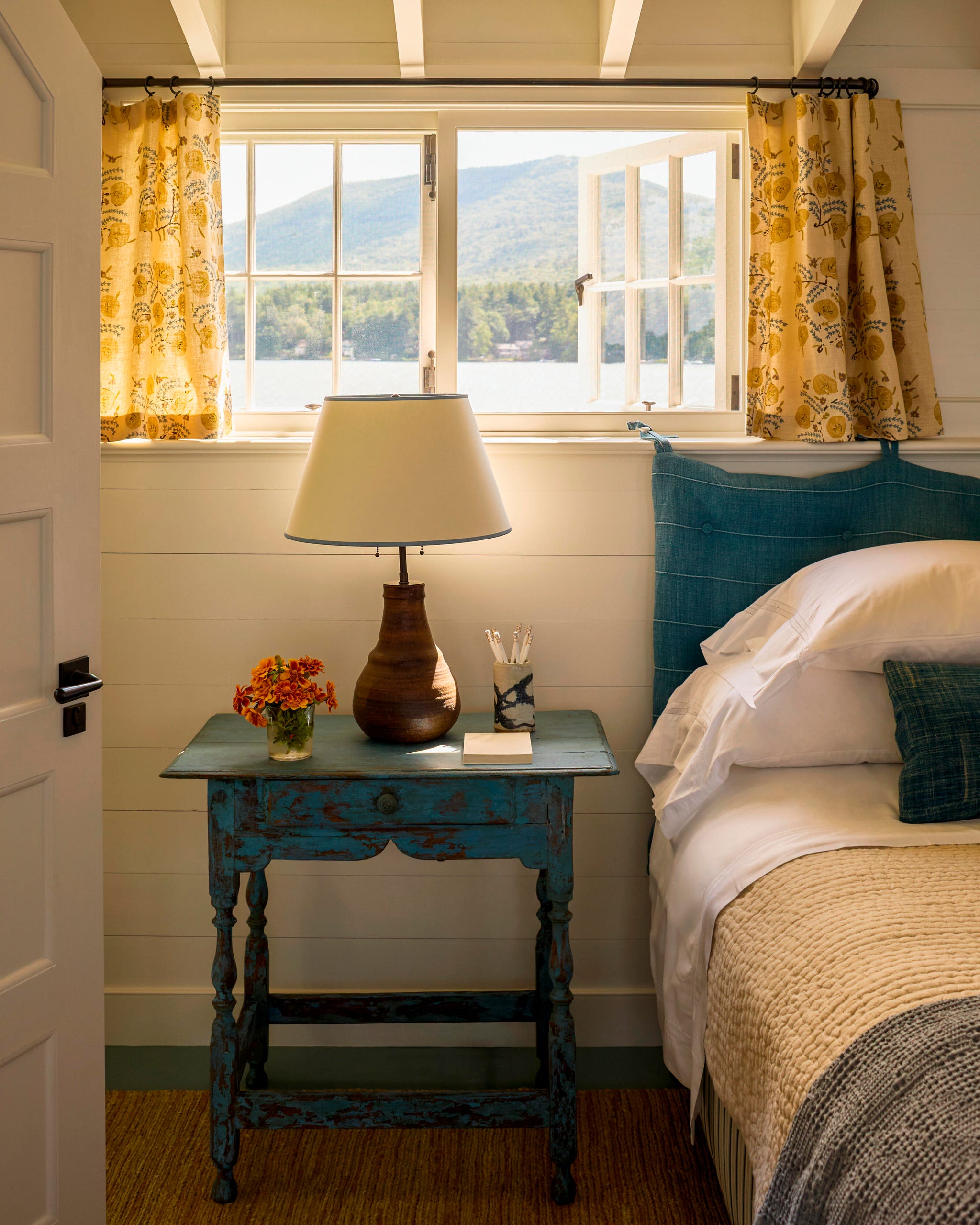 Cozy bedroom with a blue bedside table, lamp, and window view of mountains and water, featuring floral curtains and a flower vase. Cozy bedroom with a blue bedside table, lamp, and window view of mountains and water, featuring floral curtains and a flower vase.