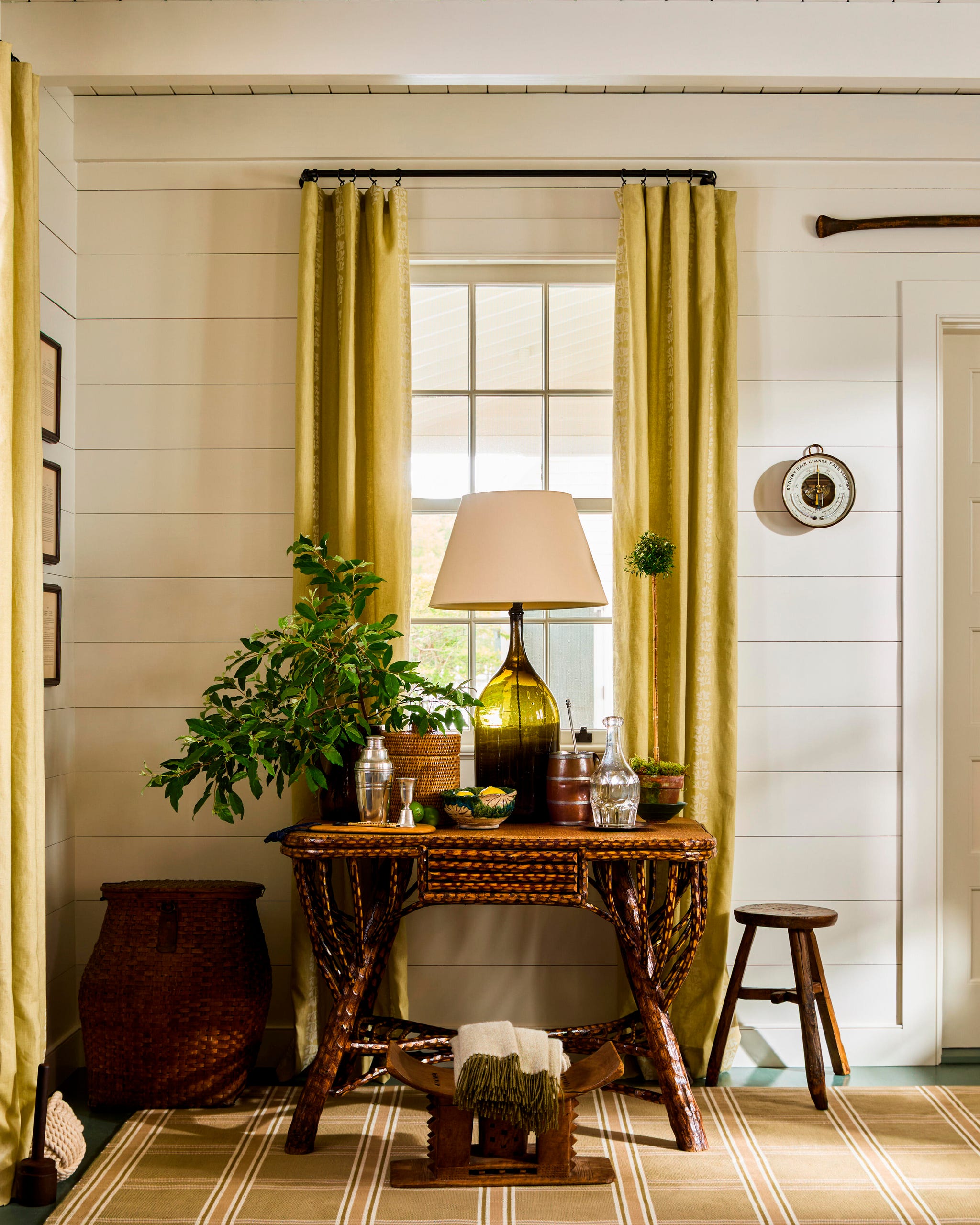 Woven table, green plants, and warm curtains, featuring a lamp and bar items on the table. Woven table, green plants, and warm curtains, featuring a lamp and bar items on the table.