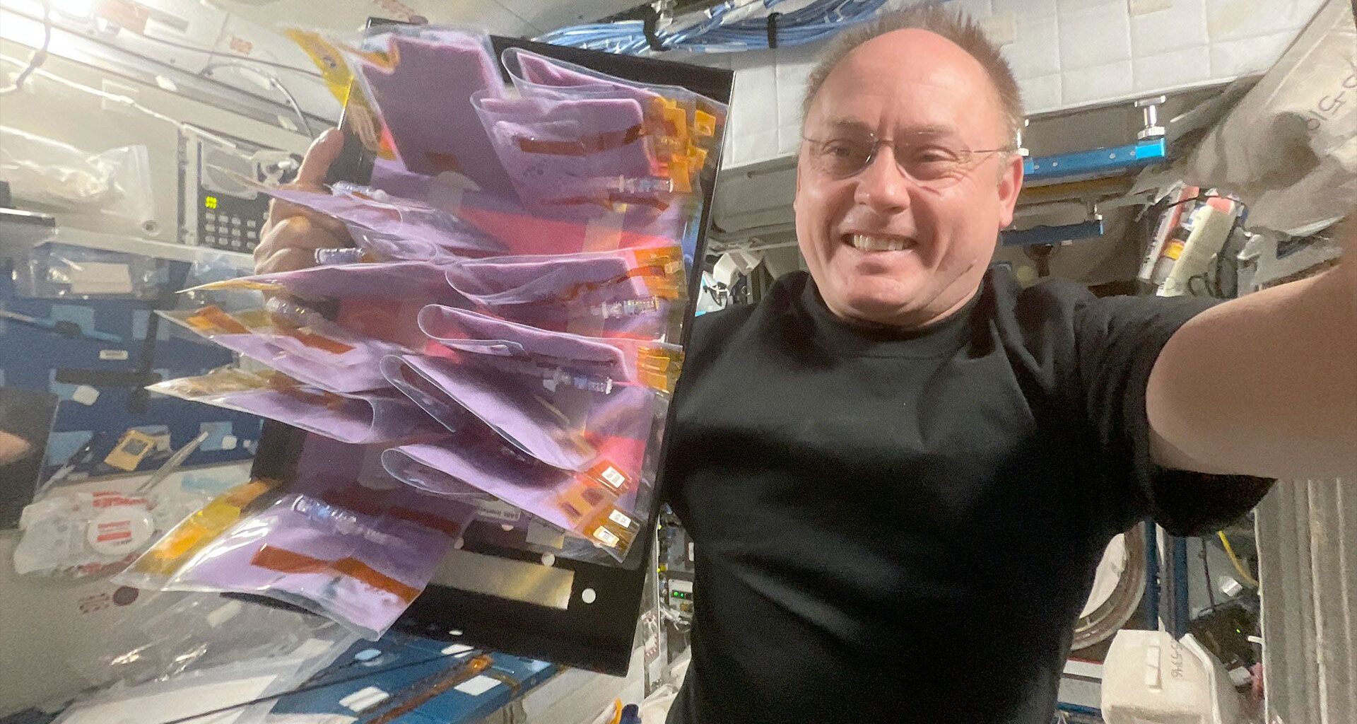 a man in a black t-shirt holds up a tray of plastic pouches filled with purple fermented milk aboard a space station.