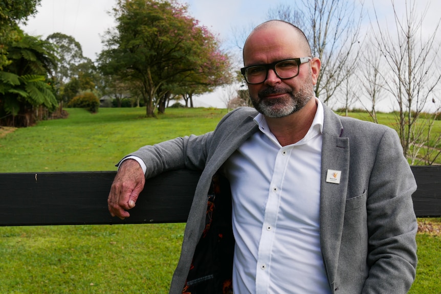 Samuel Harvey leans on wooden fence, green paddock with bushy trees behind him. 