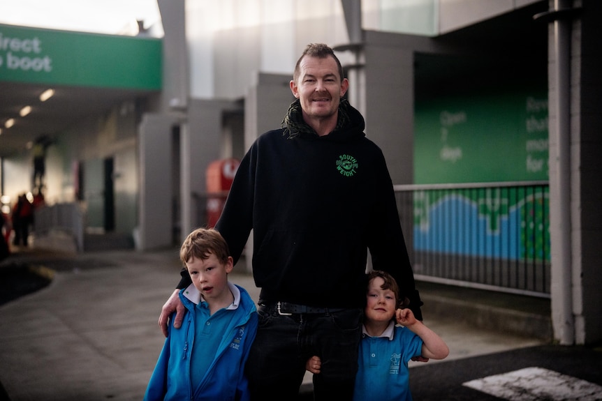 A man wearing a black hoodie stands in a carpark with his two young kids next to him wearing blue school uniform.