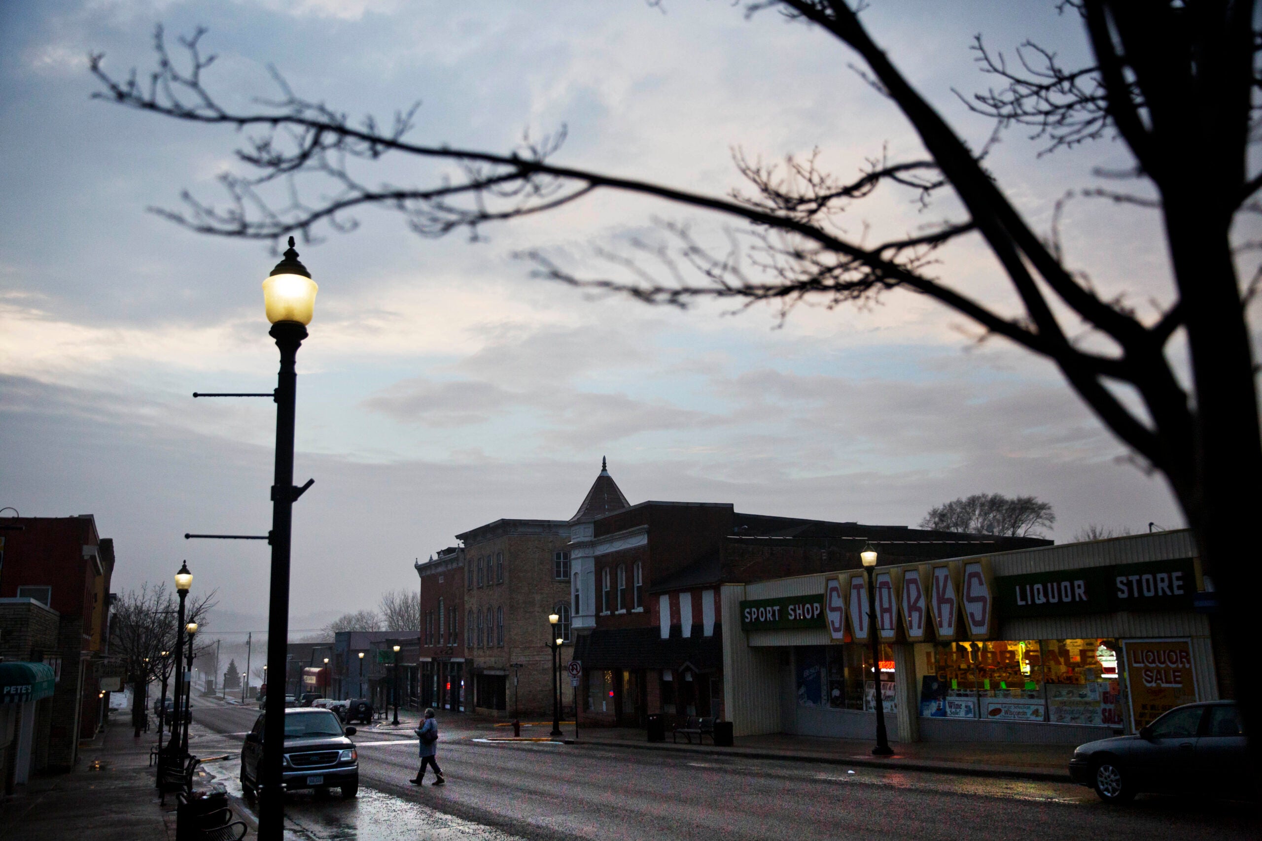 A person crosses a quiet street lined with parked cars and storefronts at dusk, with streetlights and bare tree branches visible.