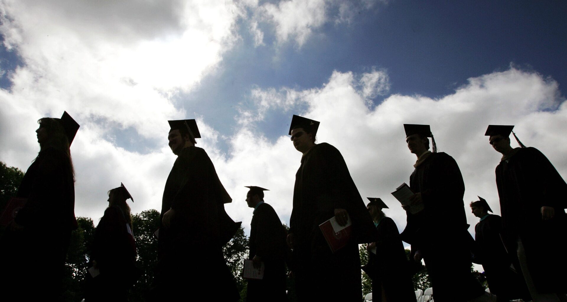 Silhouetted graduates in caps and gowns walk outdoors under a cloudy sky, holding folders, heading in the same direction.