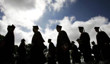 Silhouetted graduates in caps and gowns walk outdoors under a cloudy sky, holding folders, heading in the same direction.