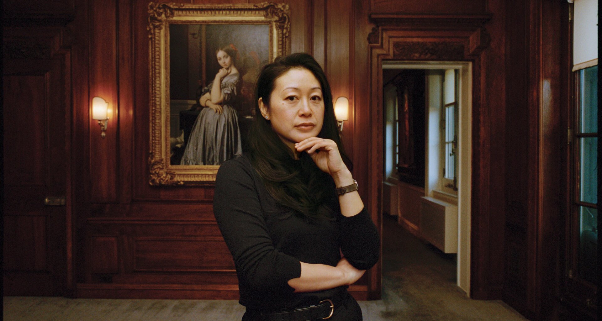 Person standing in a room with wood-paneled walls, posing thoughtfully in front of a framed portrait on the wall.