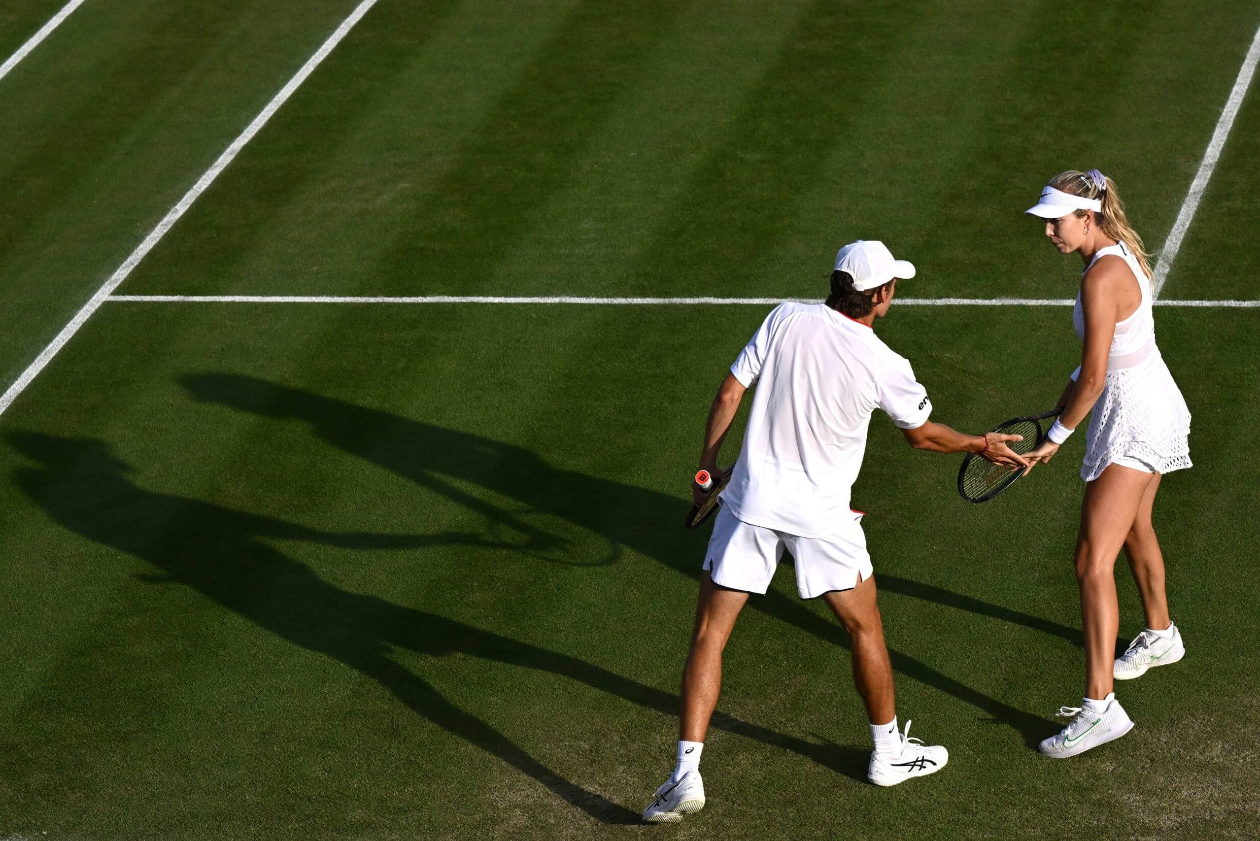 Alex de Minaur (left) and Katie Boulter (right) lo-five on a grass tennis court with their shadows stretching out to their left. They are both wearing all-white tennis outfits.