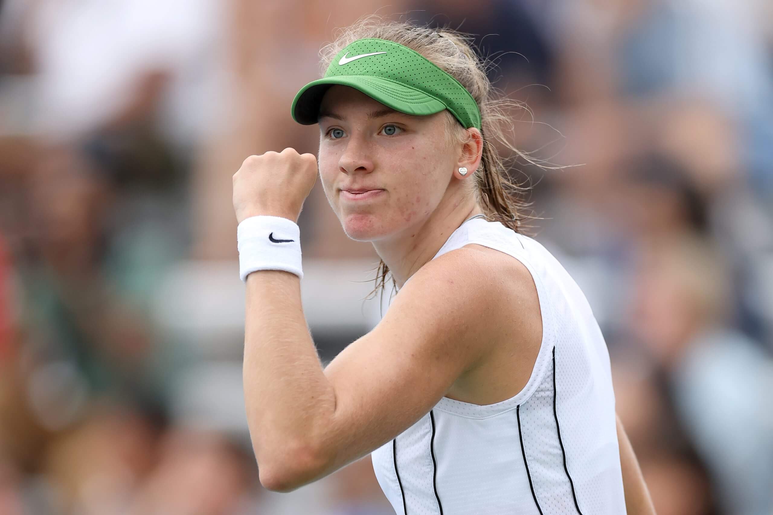 Alina Korneeva looks on pumping her left fist, wearing a white tennis outfit and green visor.