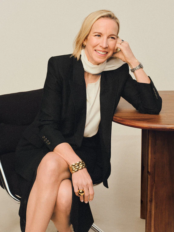 Person in a black suit sitting at a wooden table, smiling and resting head on hand.