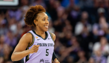 A female basketball player with curly hair, wearing a purple Golden State jersey with number 5, is running and smiling during a game.
