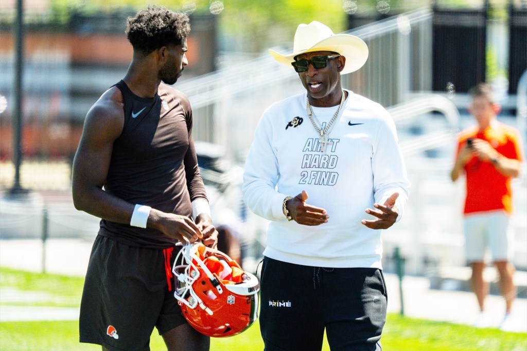 Deion Sanders in a cowboy hat and sunglasses speaks to a football player holding an orange helmet.