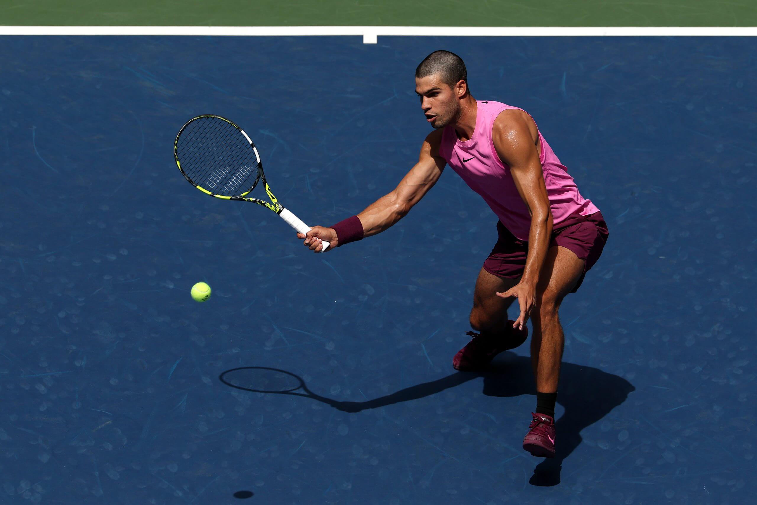 Carlos Alcaraz prepares to hit a slice volley on a blue tennis court wearing a pink-and-burgundy outfit.