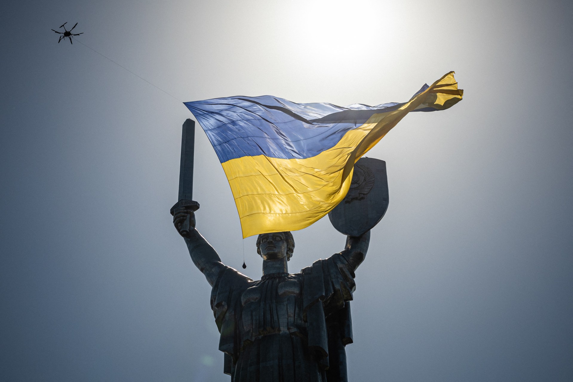 A bronze statue holding the blue and yellow Ukrainian flag aloft against a cloudy sky, with a kite visible in the upper left.