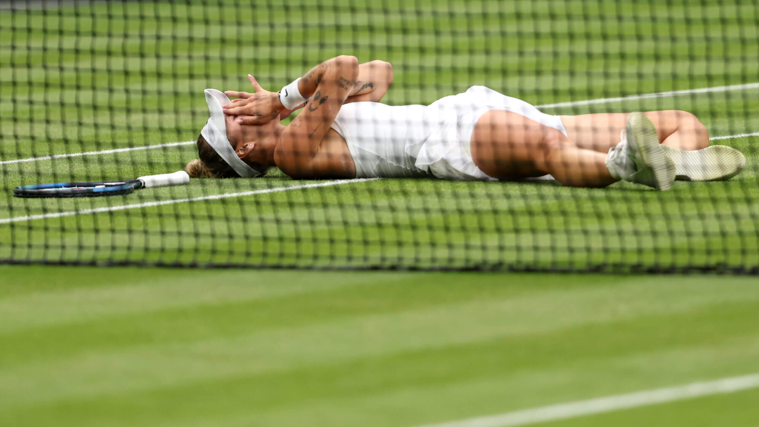 Markéta Vondroušová lies on a grass tennis court with her hands over her face in celebration.