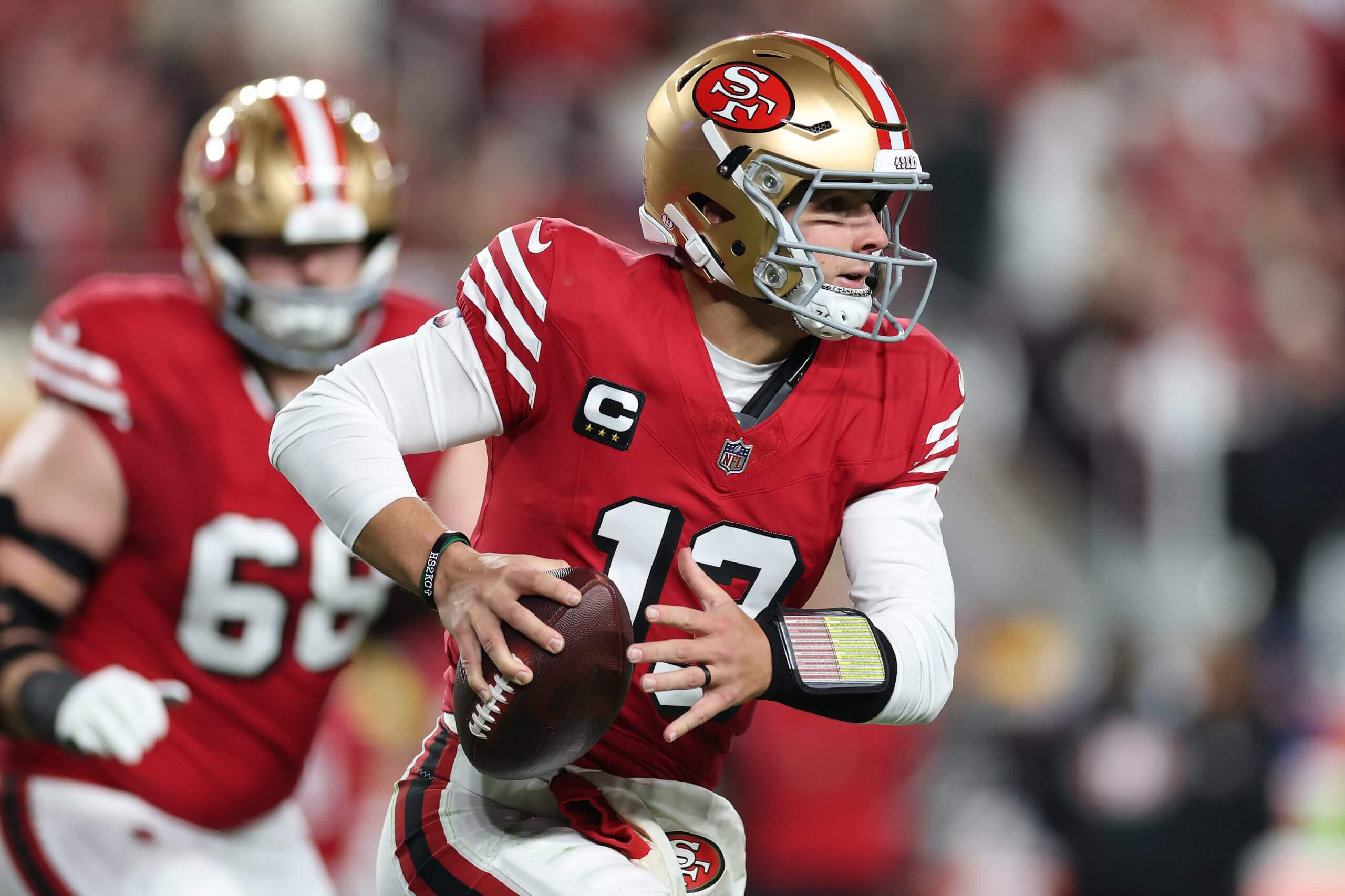 Brock Purdy, clad in a red Niners jersey and gold helmet, carries the ball in his right hand while looking to pass.
