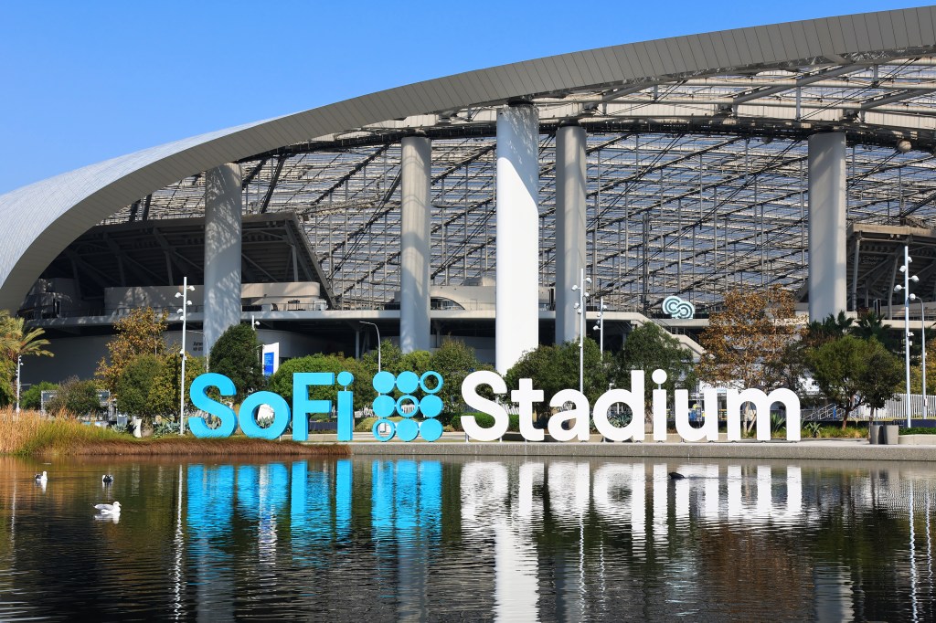 The SoFi Stadium sign in Inglewood, California, with the stadium visible in the background and its reflection in a pond.