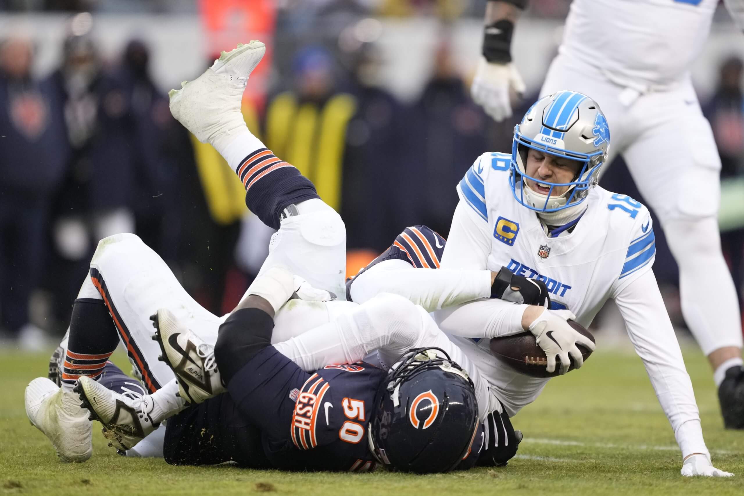 Jared Goff, clad in a white Lions uniform, is on the ground after a sack by a Chicago Bears player. He's holding the ball in his right arm and bracing himself on the ground with his left.