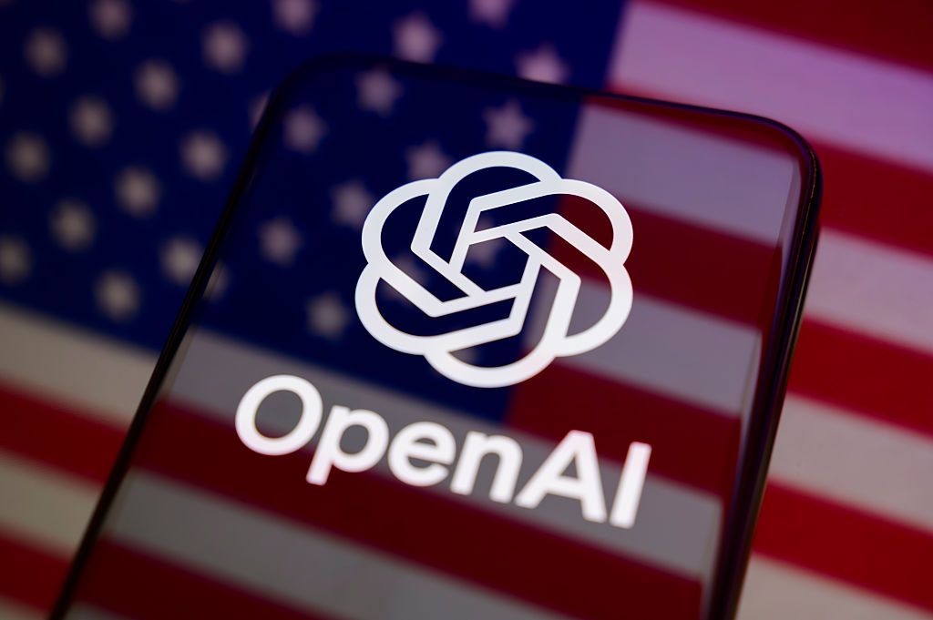 The OpenAI logo appears on the screen of a smartphone placed on a reflective surface onto which the U.S. flag is projected.