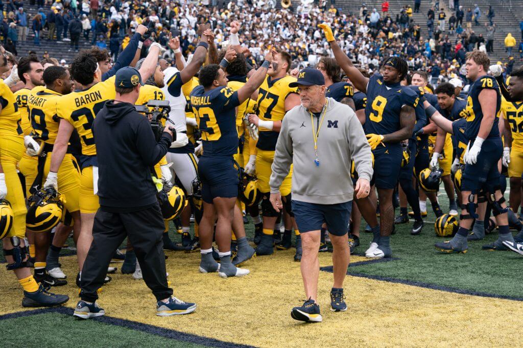 Kyle Whittingham, wearing a Michigan hat with a whistle around his neck, walks away from Michigan players huddling and raising arms on the field